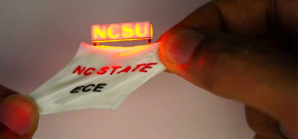 A hand holds a flexible, stretchable battery with glowing "NCSU" and "NC STATE ECE" text printed on it, highlighting its innovative design.