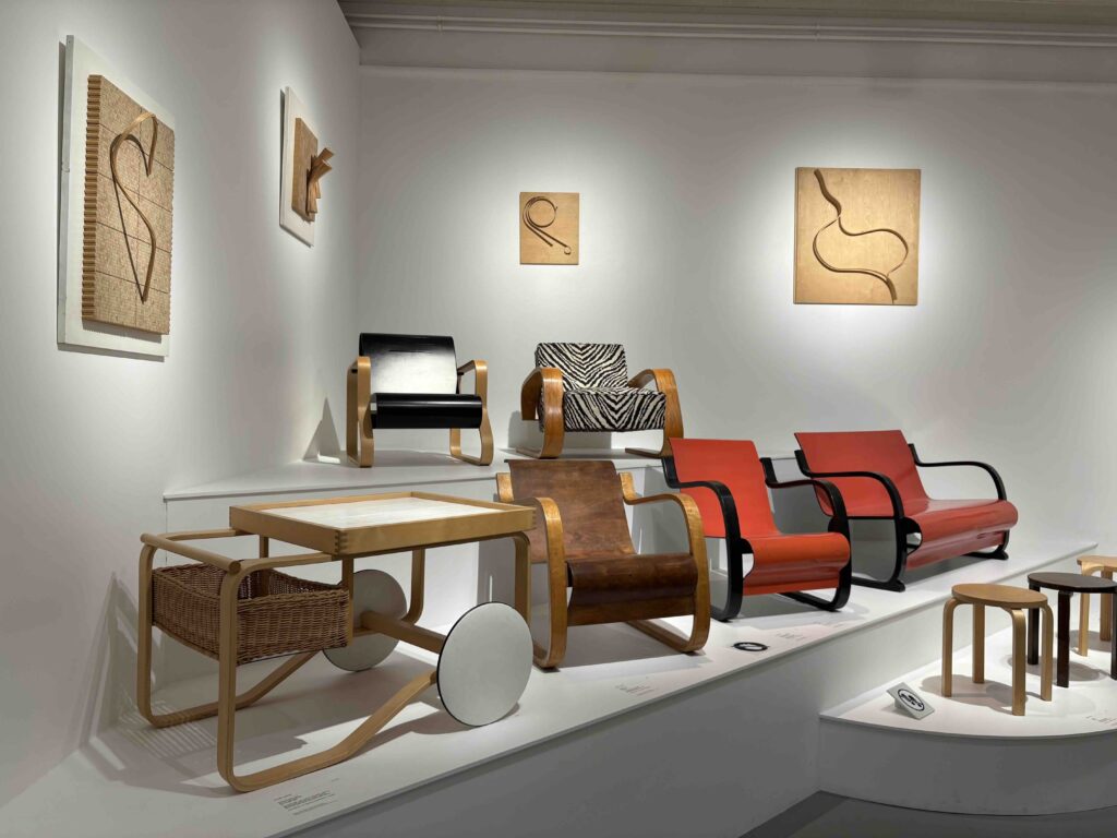 A display of modern chairs and tables atop a white shelf, featuring wood and bold colors like red and black, against a minimalistic wall.