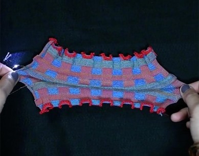 Close-up of a patterned textile swatch featuring interwoven red and blue squares, with frilled edges, displayed against a black background.