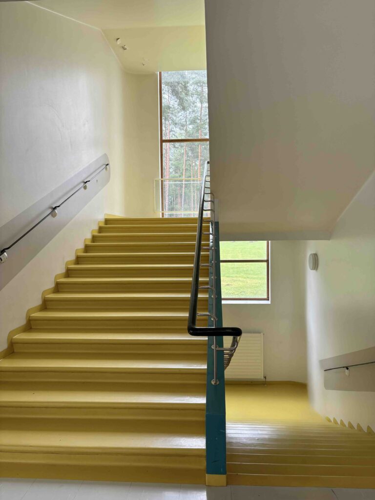 Brightly colored yellow stairs lead downwards, flanked by a white wall and a large window offering a view of greenery outside.