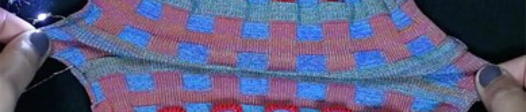 Close-up of hands holding a colorful, patterned textile with red, blue, and gray squares, highlighting the fabric's texture and design.