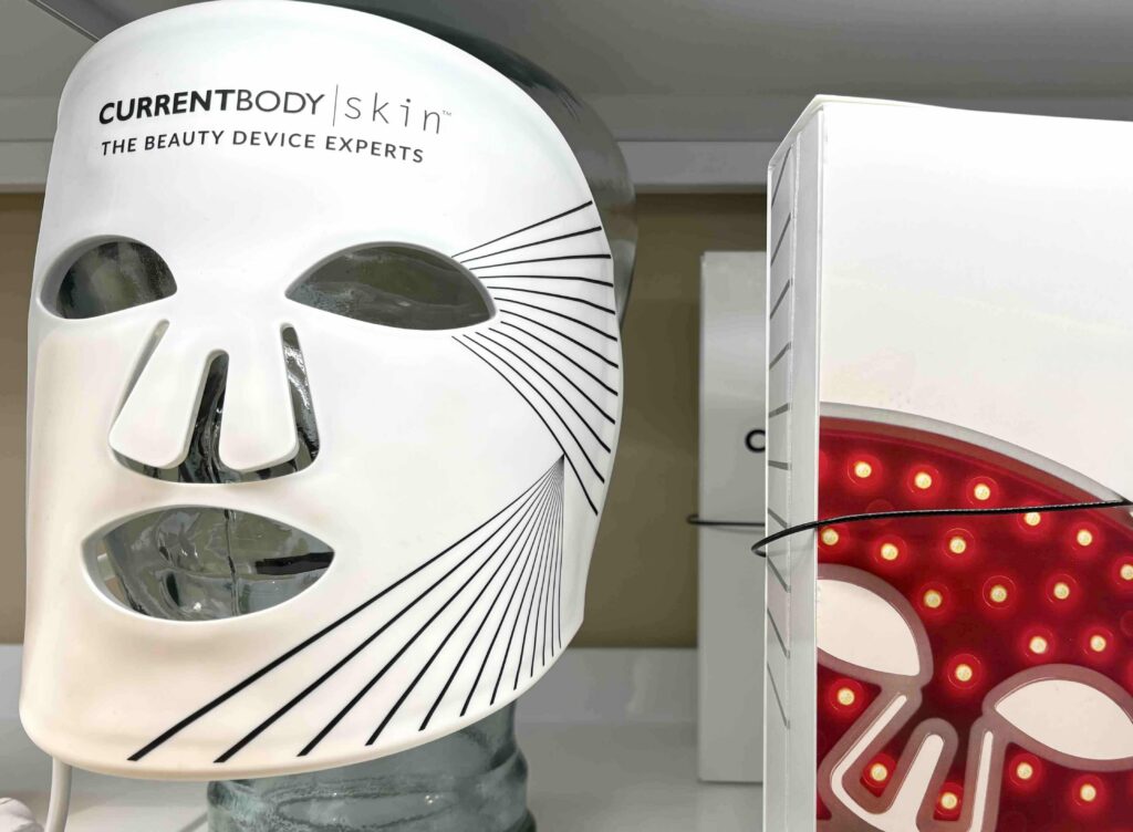 A white skincare mask with a minimalist face design, next to a red LED light device, displayed on a shelf.
