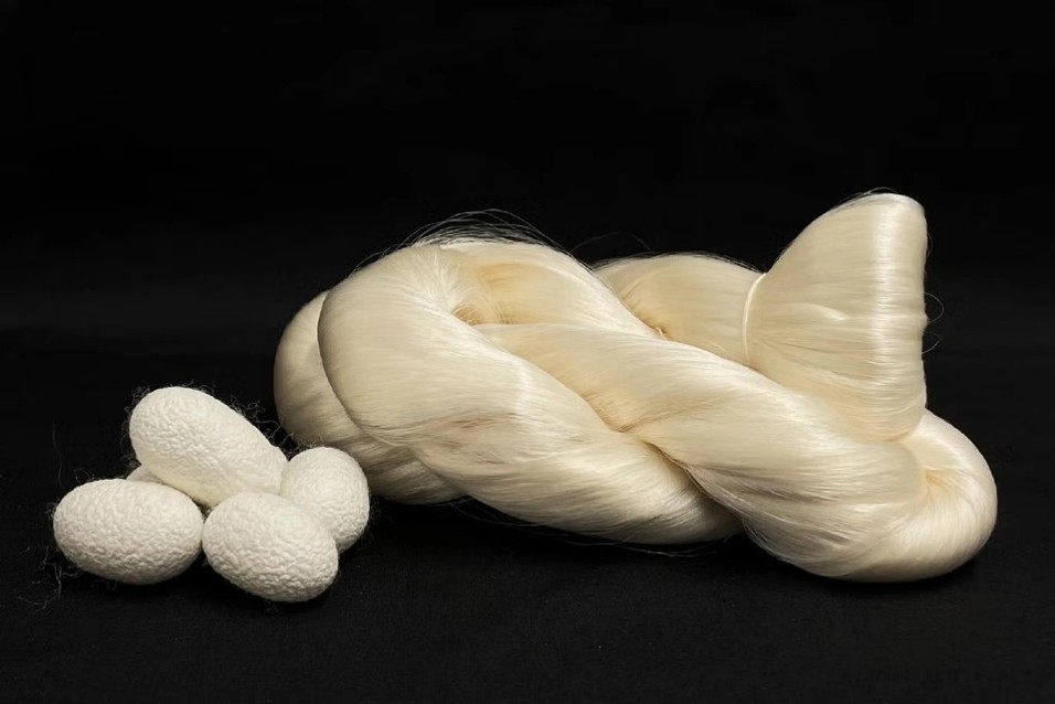 A twisted bundle of lustrous white silk fibers alongside several white silk cocoons, set against a black background.