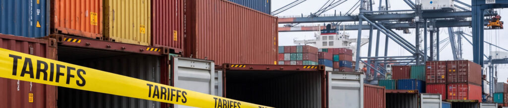 Colorful shipping containers stacked at a port, with a bright yellow "TARIFFS" banner across the foreground and cranes in the background.