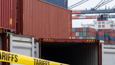 Colorful shipping containers stacked at a port, with a bright yellow "TARIFFS" banner across the foreground and cranes in the background.