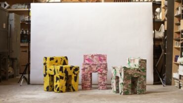 Three colorful chairs in a workshop: one yellow with black patterns, one pink, and one white with green patterns, against a plain backdrop.