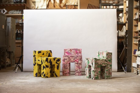 Three colorful chairs in a workshop: one yellow with black patterns, one pink, and one white with green patterns, against a plain backdrop.