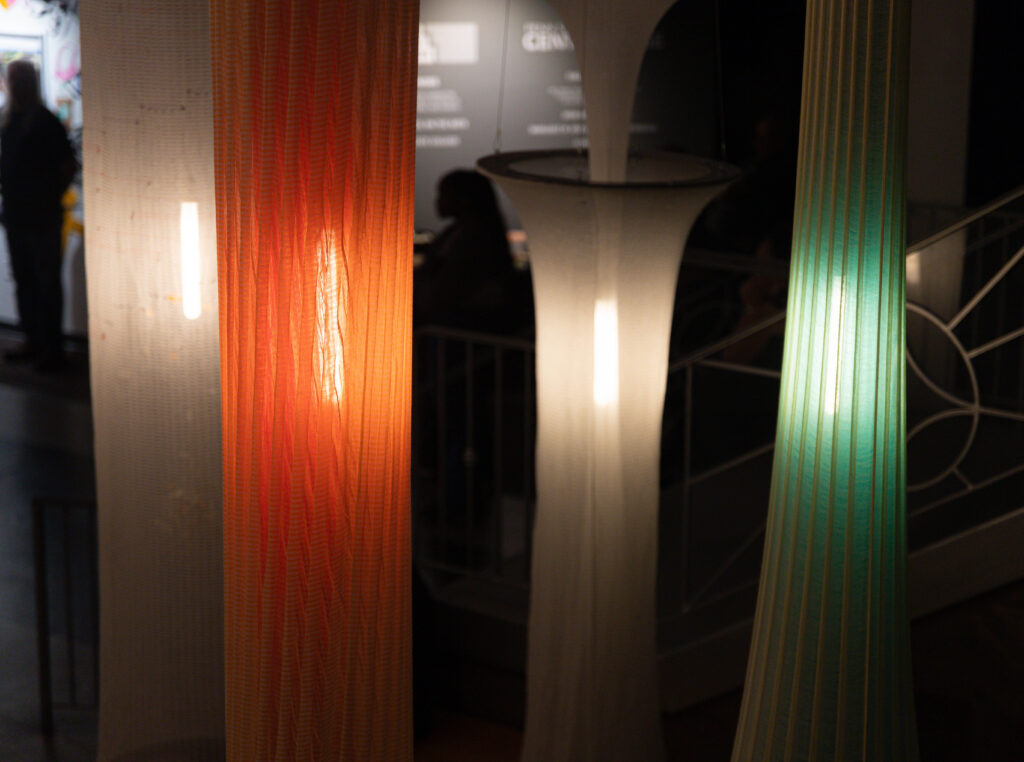 Three tall, textured light installations in orange, white, and green shades, illuminating a dimly lit space with soft glow.