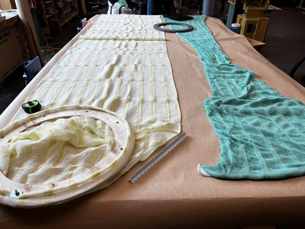 Two large knitted fabric pieces in yellow and teal drape over a work surface, alongside circular frames and measuring tools.