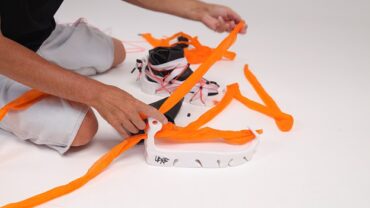 A person assembles a sneaker using bright orange pieces of material on a white surface, with a partially assembled sneaker nearby.