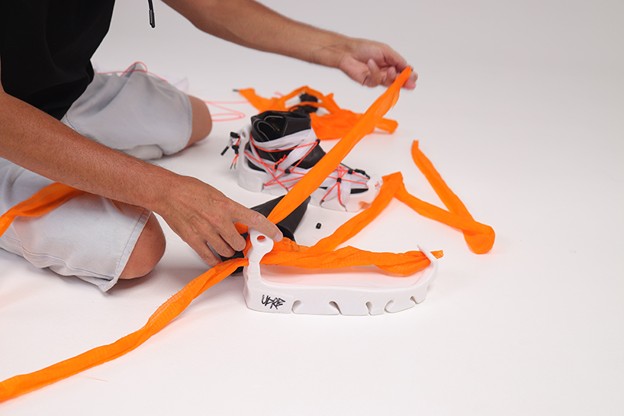 A person assembles a sneaker using bright orange pieces of material on a white surface, with a partially assembled sneaker nearby.