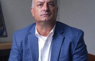 Middle-aged man with short gray hair, wearing a blue plaid blazer and white shirt, seated in an office setting with a neutral background.