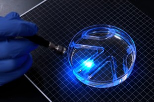 A gloved hand shines a blue light onto a transparent, circular petri dish with a geometric pattern on a grid surface.