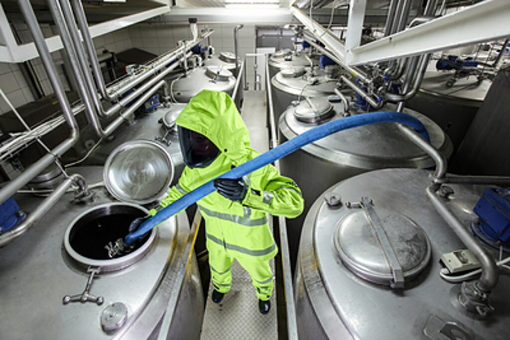A worker in a bright yellow hazmat suit uses a blue hose to transfer liquid from a large stainless steel tank in a factory setting.