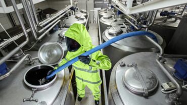 A worker in a bright yellow hazmat suit uses a blue hose to transfer liquid from a large stainless steel tank in a factory setting.