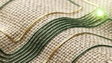 Close-up of a textured fabric with intricate, glowing green and gold conductive threads arranged in a wave pattern.