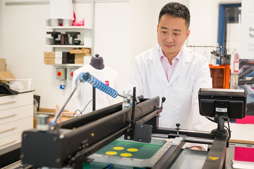 A researcher in a white lab coat operates a printing machine, focusing on a green screen with circular yellow designs in a lab setting.