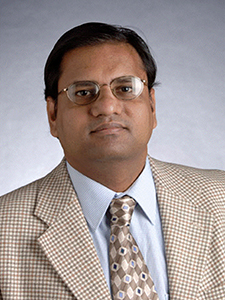 A man with glasses and a patterned tie, wearing a light-colored suit, poses against a neutral gray background.